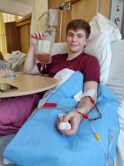 Callum Kennedy Mann pictured in hospital as he made his stem cell donation. Callum is wearing a red t-shirt and holding a bag of blood.